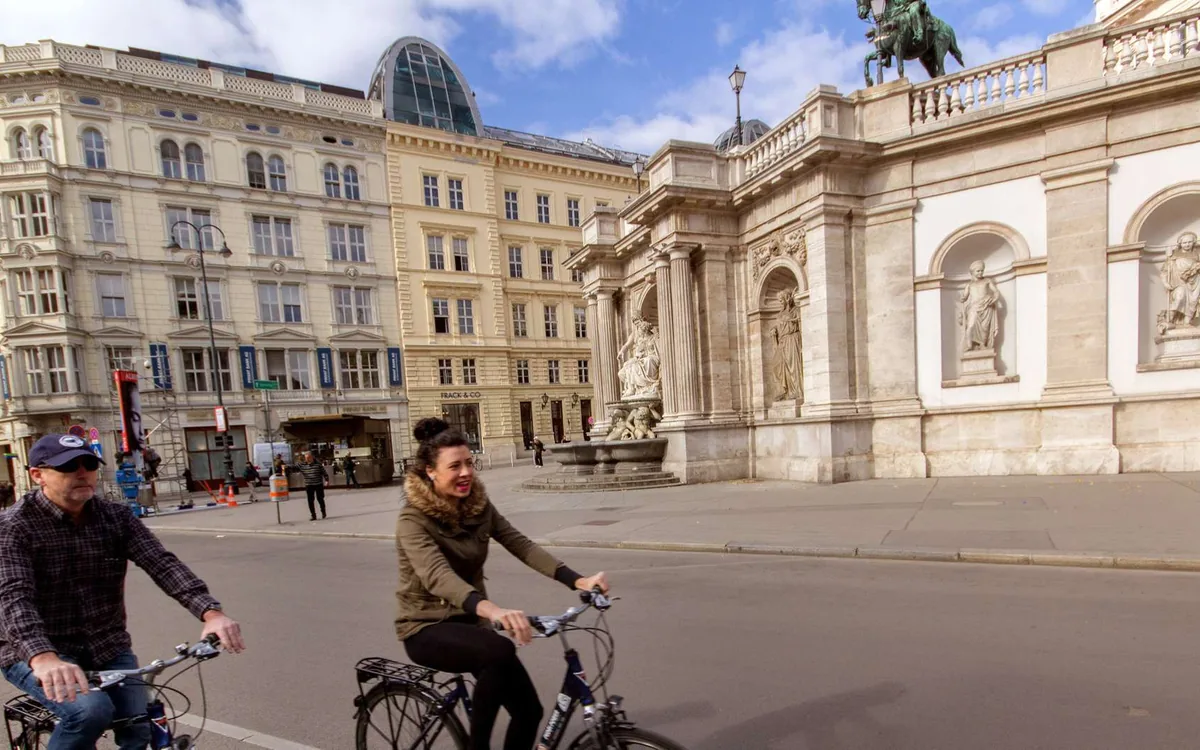 Two cyclists riding across a wide square past grand stone buildings in Vienna