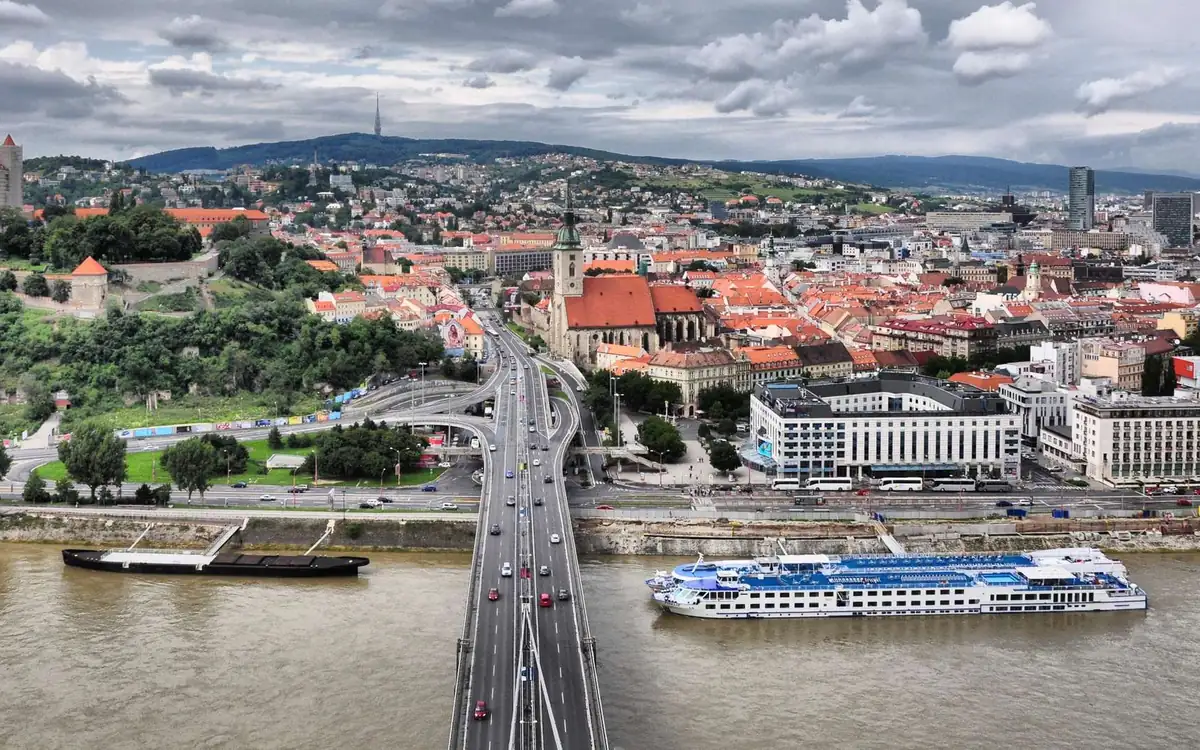 Bratislava old town roofs and a cathedral tower above the river, with a road bridge and a river ship