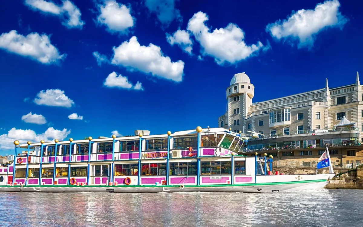 A white riverboat with pink trim on the water, a domed building on the bank behind