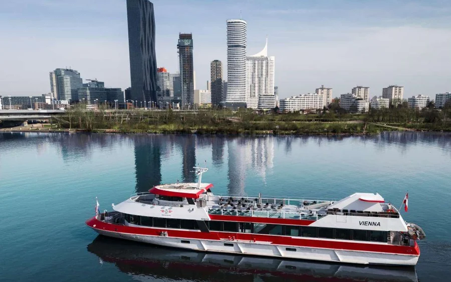 A red and white river boat on the Danube below city towers