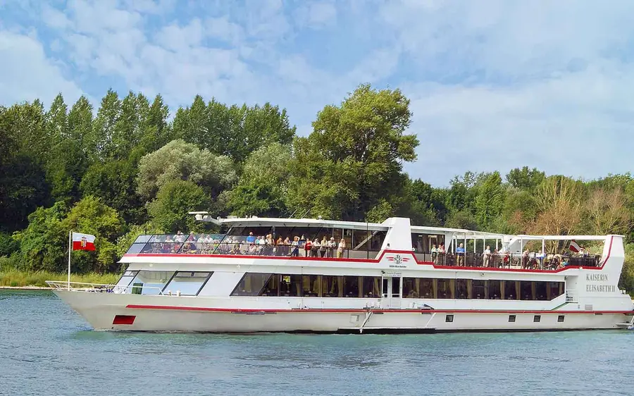 A white river cruise boat passing a wooded riverbank