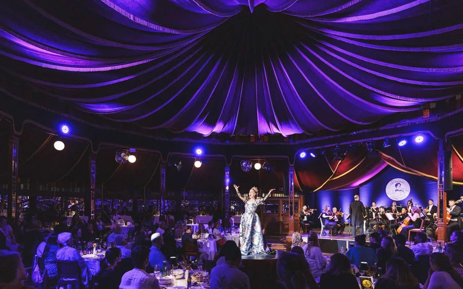 A singer on stage before dinner tables under a purple-lit tent roof